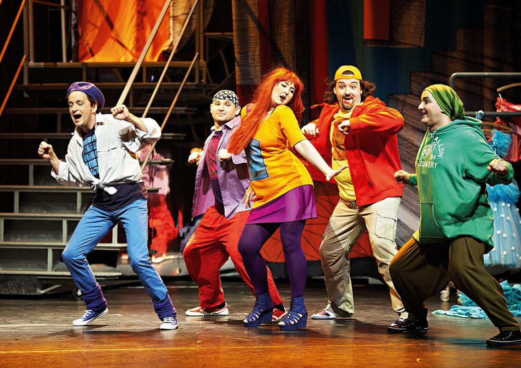 A group scene, the five dancing soloists on the stage, in a pose with slightly bent legs and hands folded in fist, all in colourful, youthful costumes and headgear. The first character of a man in blue narrow trousers, a light shirt tied at the waist and 