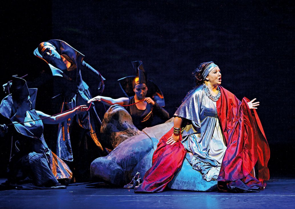 A group scene, in the foreground, a soloist in a stylized antique silver dress, red cloak and a silver hairband, sits on a large male sculpture. Behind her in dark light three figures in long, shiny dresses with large, stiff collars and geometric headgear