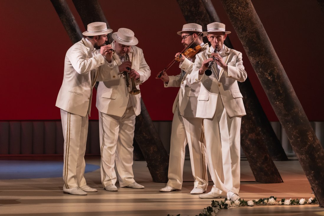 Czterech mężczyzn w białych strojach z kapeluszami na głowach i z instrumentami w ręku wykonujący muzykę.