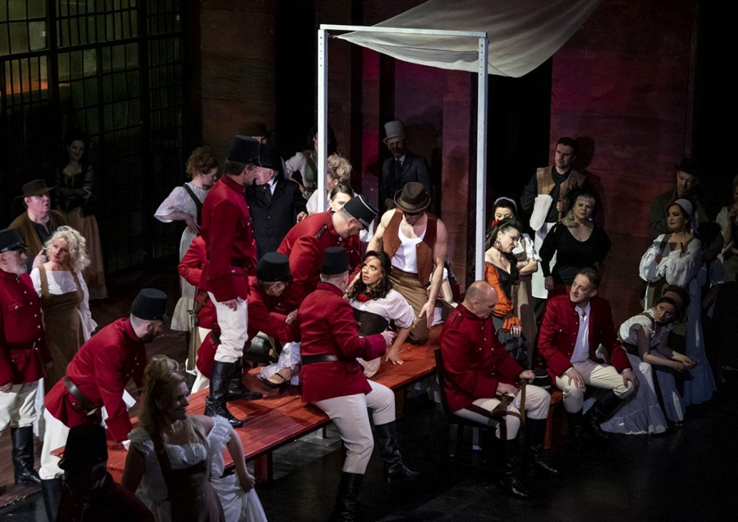 A group scene, in the central point on a long wooden bench, the soloist in a white blouse with a red rose pin and in a dark corset sits leaning backwards leaning on bent hands. She is surrounded by men in guards' outfits - white trousers put on high boots