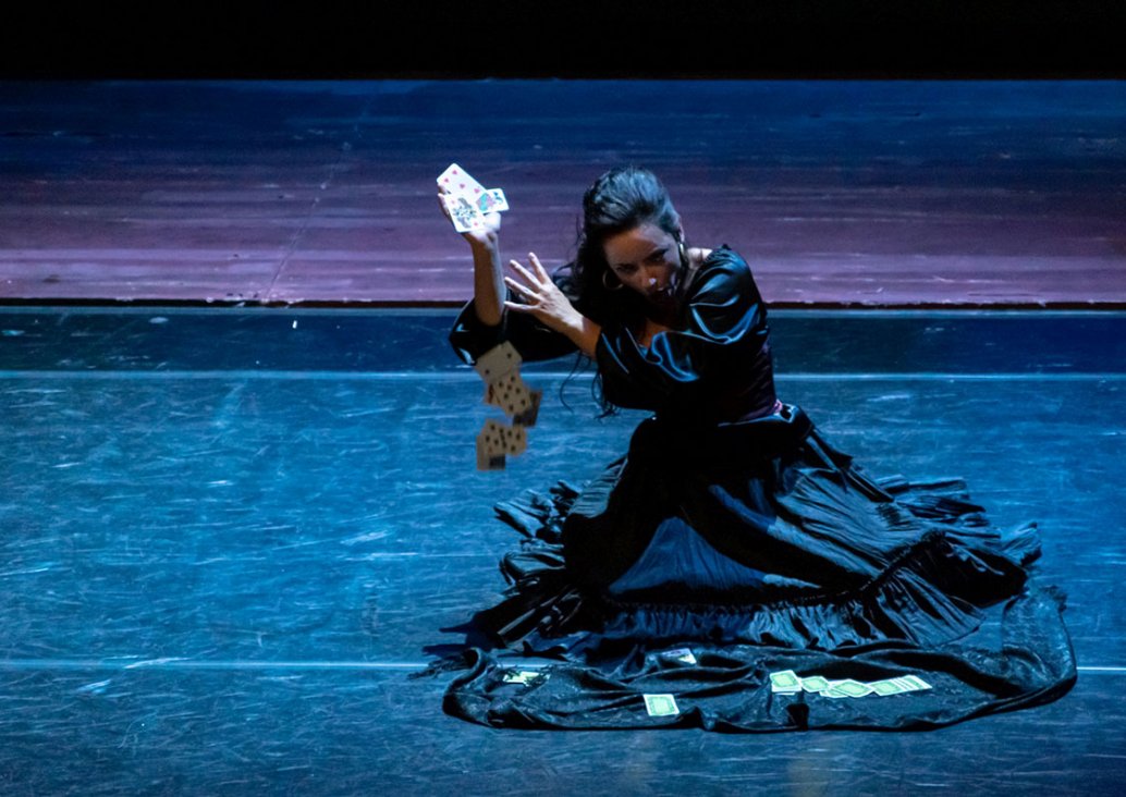 A solo scene, in the middle of the stage, kneels down a soloist in a shiny black Spanish dress. In her right raised hand she holds three cards, the others fall to the ground, some lie on her dress.