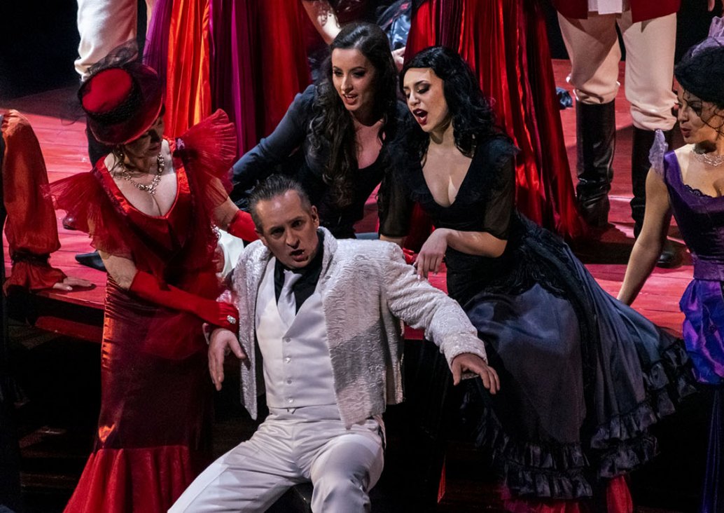 A group scene,  in the middle a soloist in white trousers, a white vest, a black shirt, a white tie and a white, shiny short jacket without buttons sits on a chair. Behind him, on a large, round, wooden table, there are two soloists in black, flouncy Span