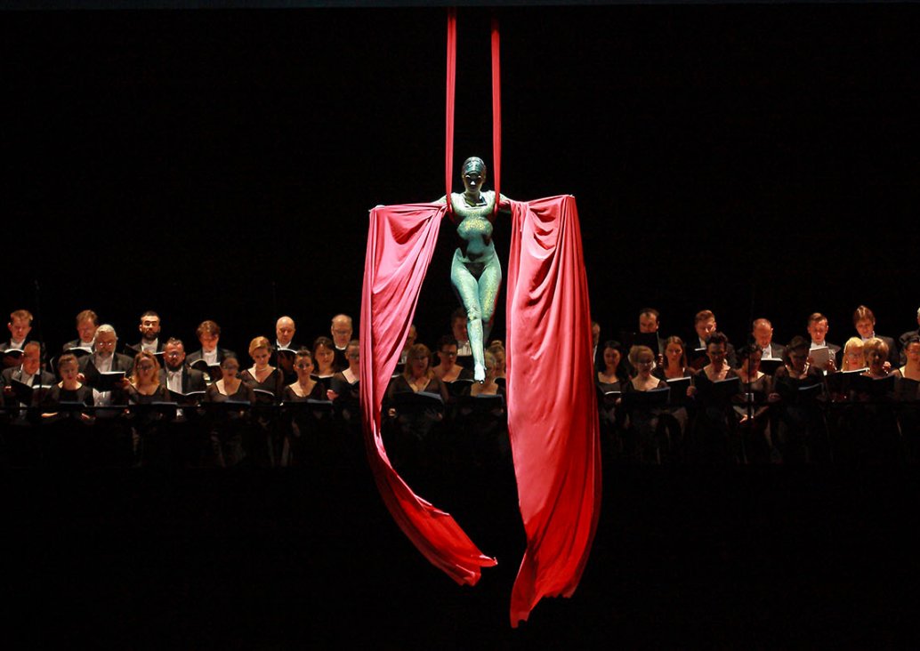 A group scene, in the middle of the stage in the air an acrobat in a shiny, smooth outfit adjacent to the whole body. She is held on a red sash, with her legs lowered - right leg is slightly forwarded. In both hands, unfolded to the sides, she holds long 