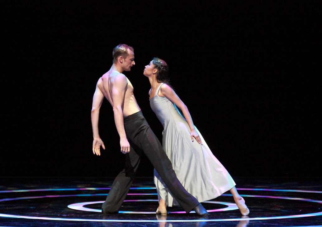 A scene involving two dancers. A man in long, black trousers, without a shirt, leaning back, with his hands lowered and his right leg straightened forward. A woman in a white, smooth dress on shoulder straps, leaning towards the man, with her left leg str