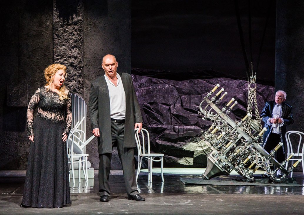 A duet scene with two soloists, a woman in a black long lace dress, a man in a black suit with a long jacket and a white shirt, stand next to each other. On the stage several white chairs, a large chandelier on chains on the floor, in the background walls