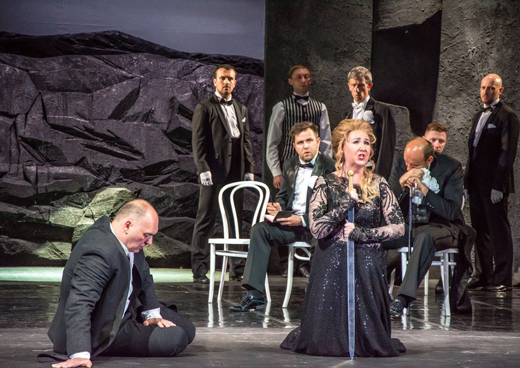 A group scene, in the foreground two soloists. A man in a black suit is sitting on the floor supported by his right hand, a woman in a black lace dress is kneeling and holding a sword facing the floor in her hands. In the background, men in dark, evening