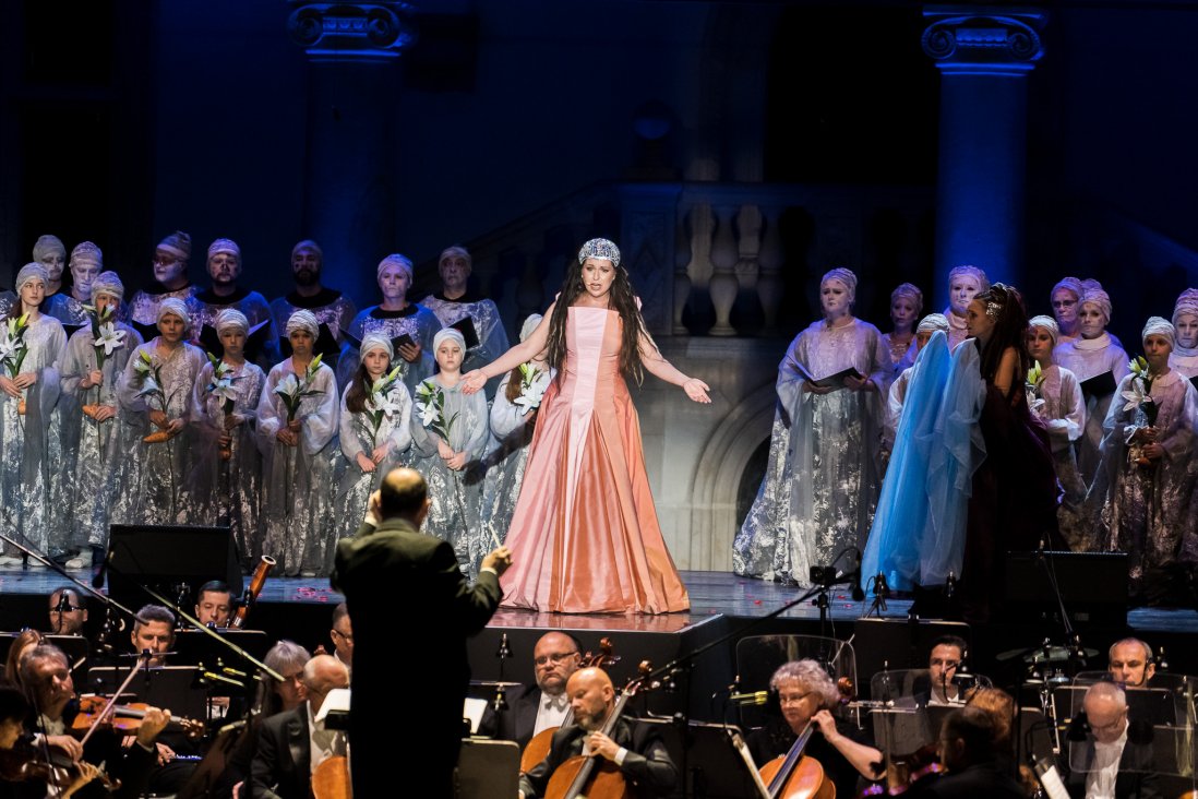 Wanda w różowej sukni i srebrnym diademie rozkłada ręce w dramatycznym geście. Wokół niej chór i przerażona Panna trzymająca woal. Na pierwszym planie dyrygent i orkiestra.