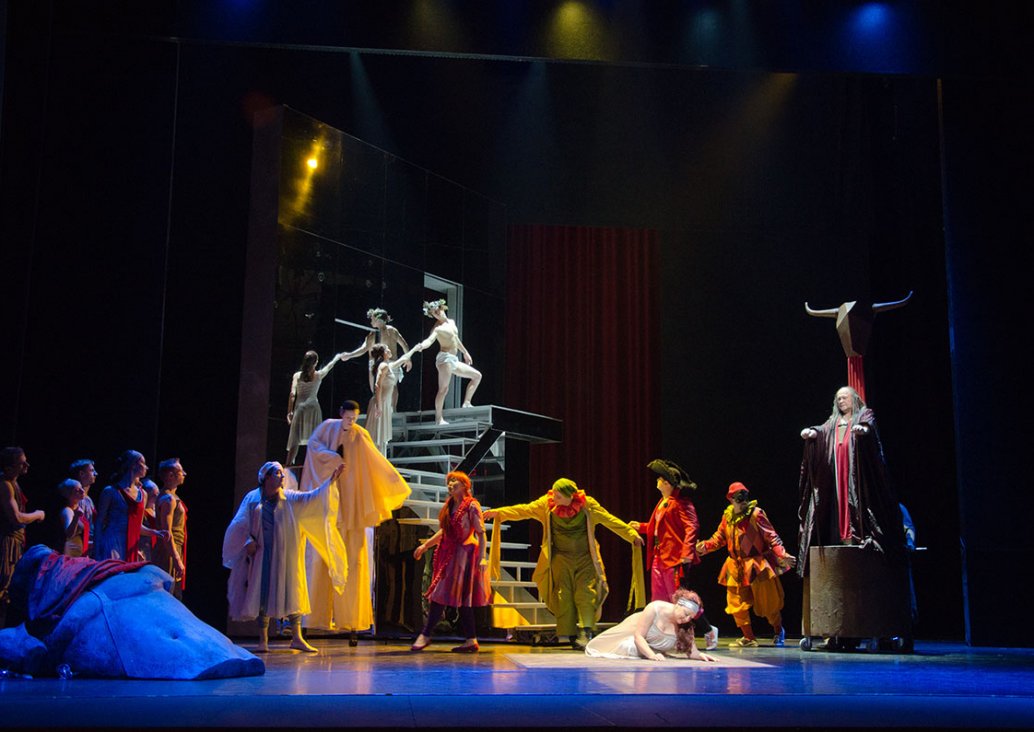 Scena zbiorowa, w centralnym punkcie solistka w srebrnej sukni na ramiączkach siedzi pochylona na ziemi i podpiera się rękami. Po lewej stronie rzeźba mężczyzny, za nią grupa postaci w luźnych brązowo-czerwonych strojach, obok niech postaci w długich, zwi