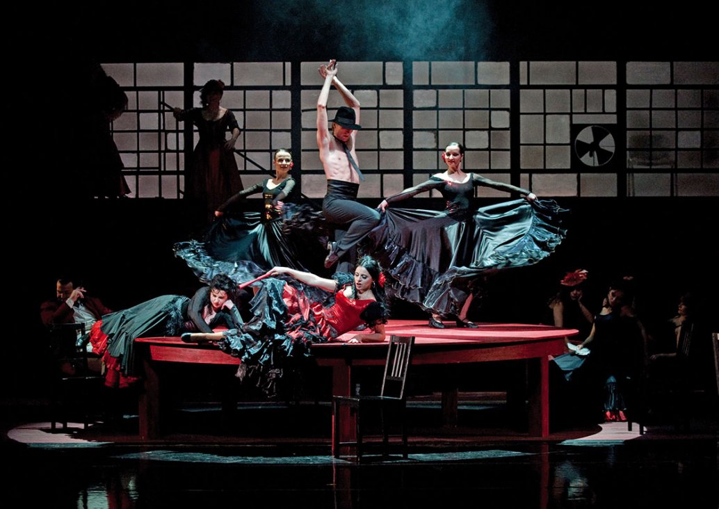 Scena zbiorowa, na dużym, okrągłym, drewnianym stole dwie kobiety w hiszpańskich, czarno-czerwonych, falbaniastych sukniach leżą na boku podparte na rękach. Za nimi troje tancerzy - dwie kobiety w hiszpańskich, czarnych, falbaniastych sukniach, trzymają w