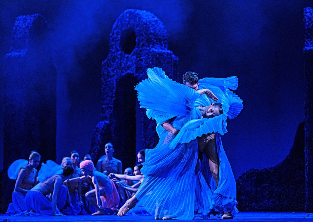 A group scene, in the foreground a couple of dancers, a woman in a pleated blue dress in a backward curved pose, supported by a man. In the background, characters sitting on the floor and geometric decorations.