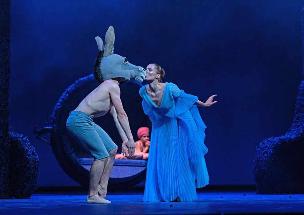 A duet scene with a couple of dancers. A man without a T-shirt, in blue bermuda, with a donkey head, stands on bent knees with his hands down. A woman in an airy, pleated blue dress kisses him leaning towards him. In the background, a boy in a pink turban