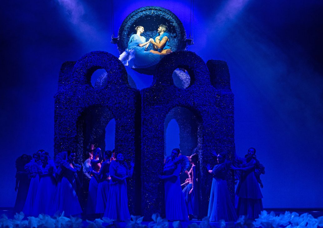 A group scene using large size decorations with geometric shapes and blue light. A large group of characters in airy, blue, pleated dresses gathered around the decoration. Above them, in a circle hanging over the stage, a couple sits, holding hands and lo
