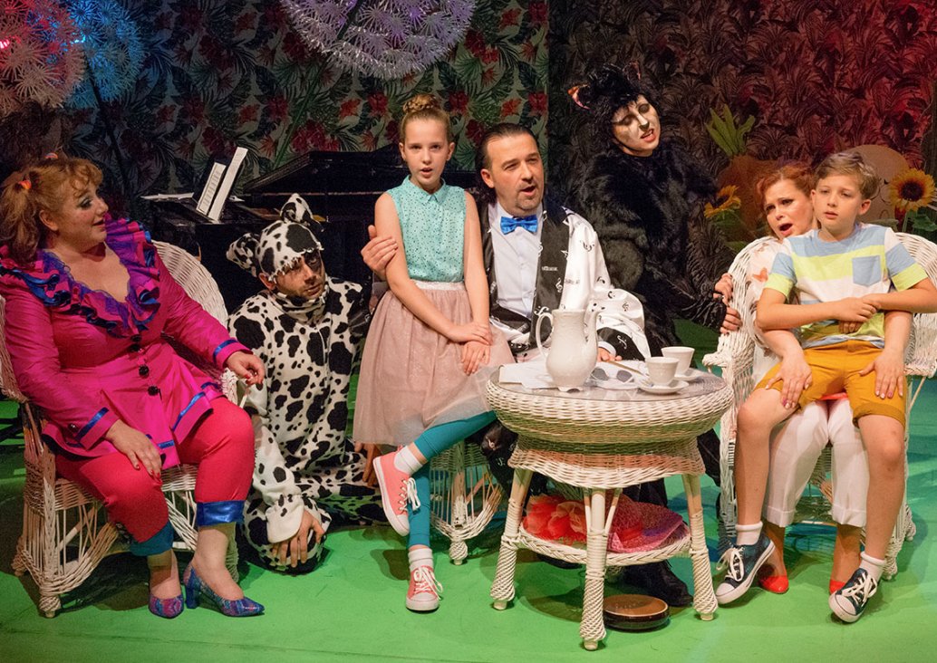 A group scene. Three actors in colourful costumes sit on wicker armchairs. There are two children on their knees. Between them there are actors dressed as dog and cat. On the wicker table there is a teapot and two cups. A piano stands in the background.
