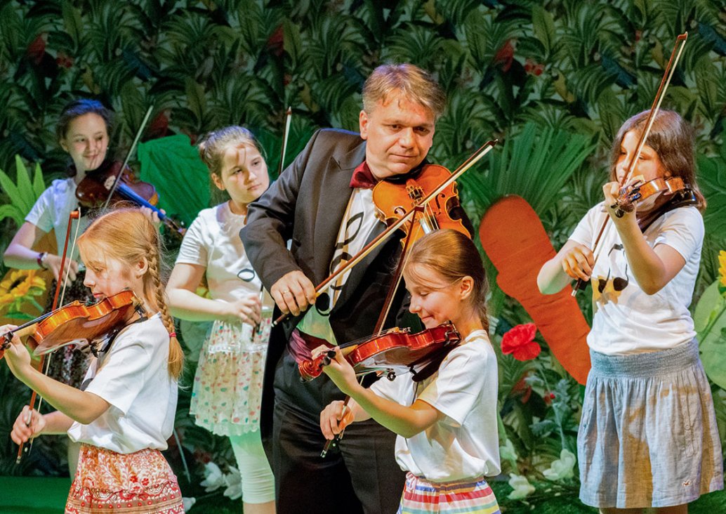 A group scene. An instrumentalist playing the violin in the middle, five girls around him, dressed in bright T-shirts and skirts, also playing the violin.