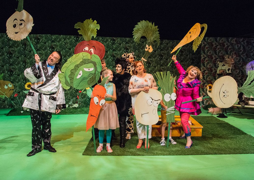 A group scene with adults and two children. All dressed in colorful costumes, two actors dressed as cat and dog. Everybody is holding large images of popular vegetables made of cartons in their hands.