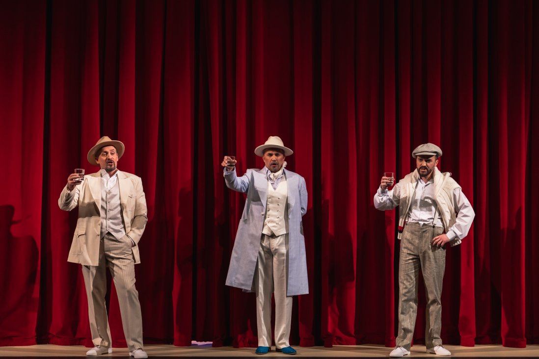 Trzech śpiewających bohaterów, w centrum Don Alfonso, eleganckich strojach i kapeluszach na tle spuszczonej kurtyny z kieliszkami w dłoniach uniesionymi na znak toastu.