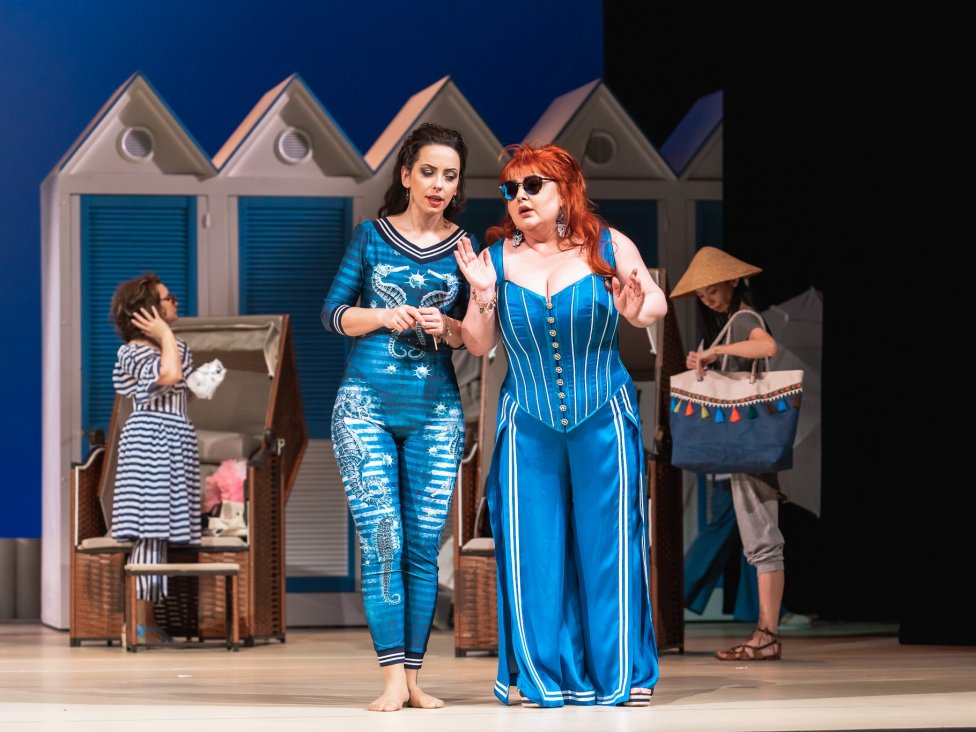 Dwie kobiety, Dorabella i Fiordiligi) w niebieskich strojach śpiewające na scenie. W tle plażowicze i biało-niebieskie parawany. Scenografia przypomina plażę.
