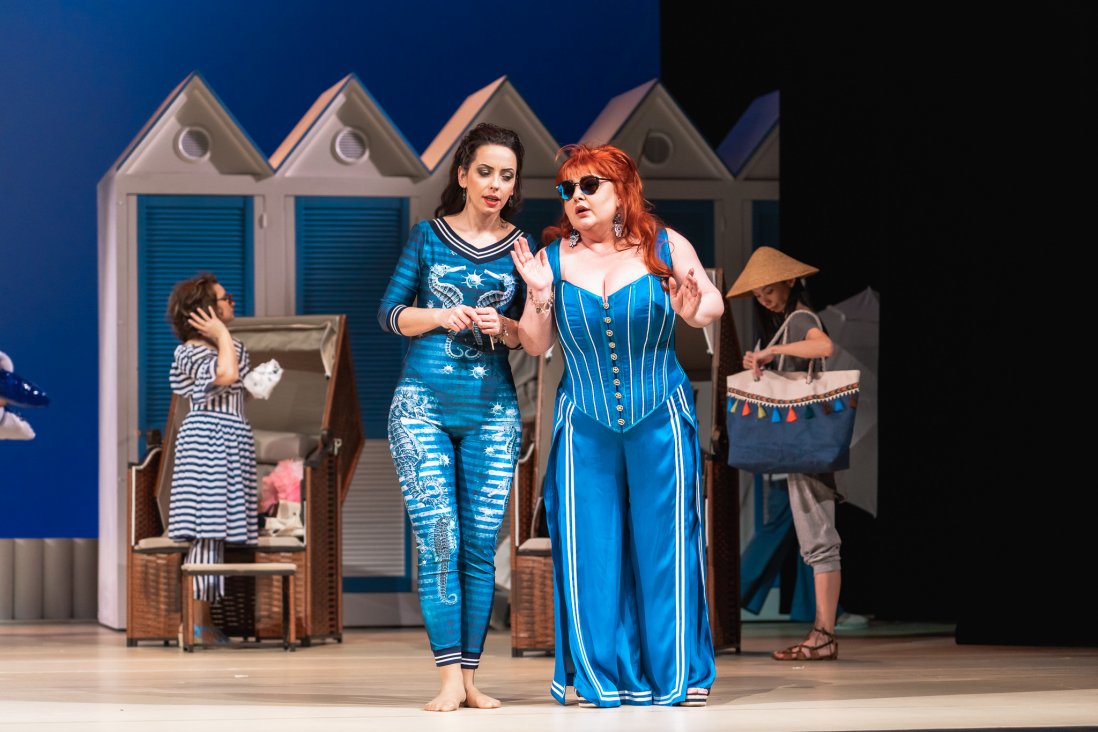 Dwie kobiety, Dorabella i Fiordiligi) w niebieskich strojach śpiewające na scenie. W tle plażowicze i biało-niebieskie parawany. Scenografia przypomina plażę.
