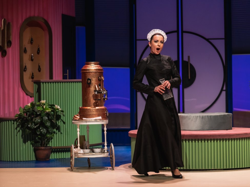 Śpiewająca służąca Despina na tle błękitno-zielono-różowej scenografii przypominającej wczasowy pensjonat.