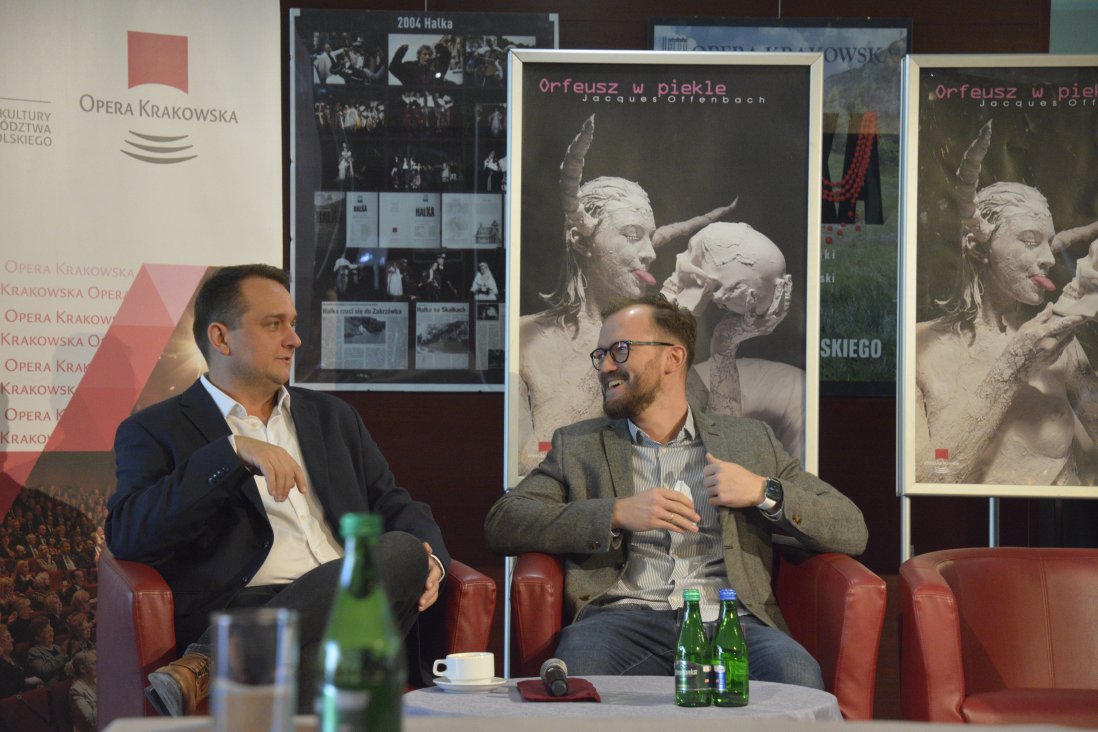 Dominik Sutowicz i Jarosław Bielecki siedzą w czerwonych fotelach, uśmiechają się do siebie. W tle plakaty spektaklu.