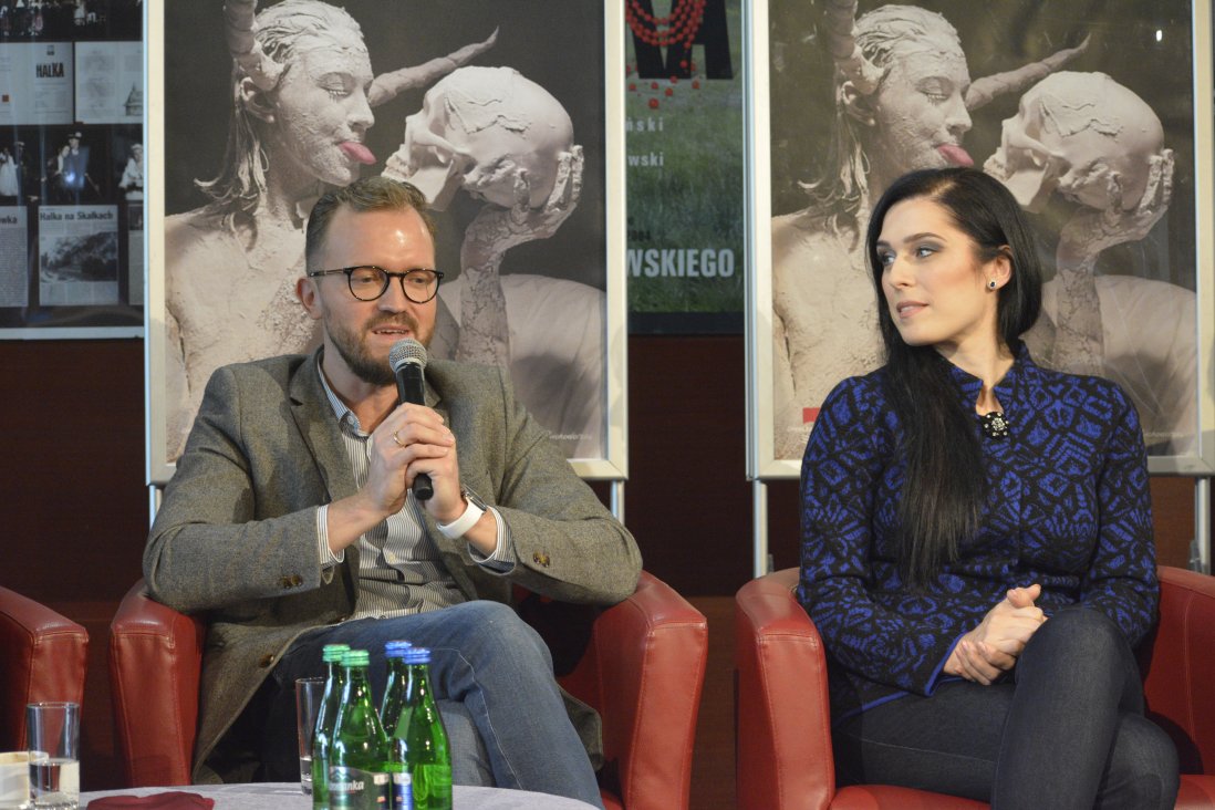 Jarosław Bielecki (Orfeusz) wypowiada się do mikrofonu. Po prawej Marcelina Beucher (Eurydyka), patrzy w jego stronę. W tle plakaty spektaklu.
