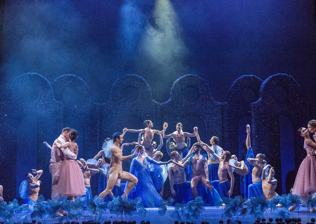 A group scene, dancers in different arrangements all over the stage. On opposite sides, two pairs in embrace, through the center runs the main man character dressed only in bodily underwear. In the middle of the stage, a group of dancers in different pose