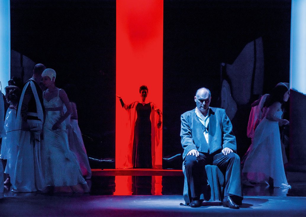 A group scene, in the foreground the soloist in a suit sits on a chair covered with fabric, on the sides characetrs in motion in white dresses, in the background the soloist stands in a red light rectangle.