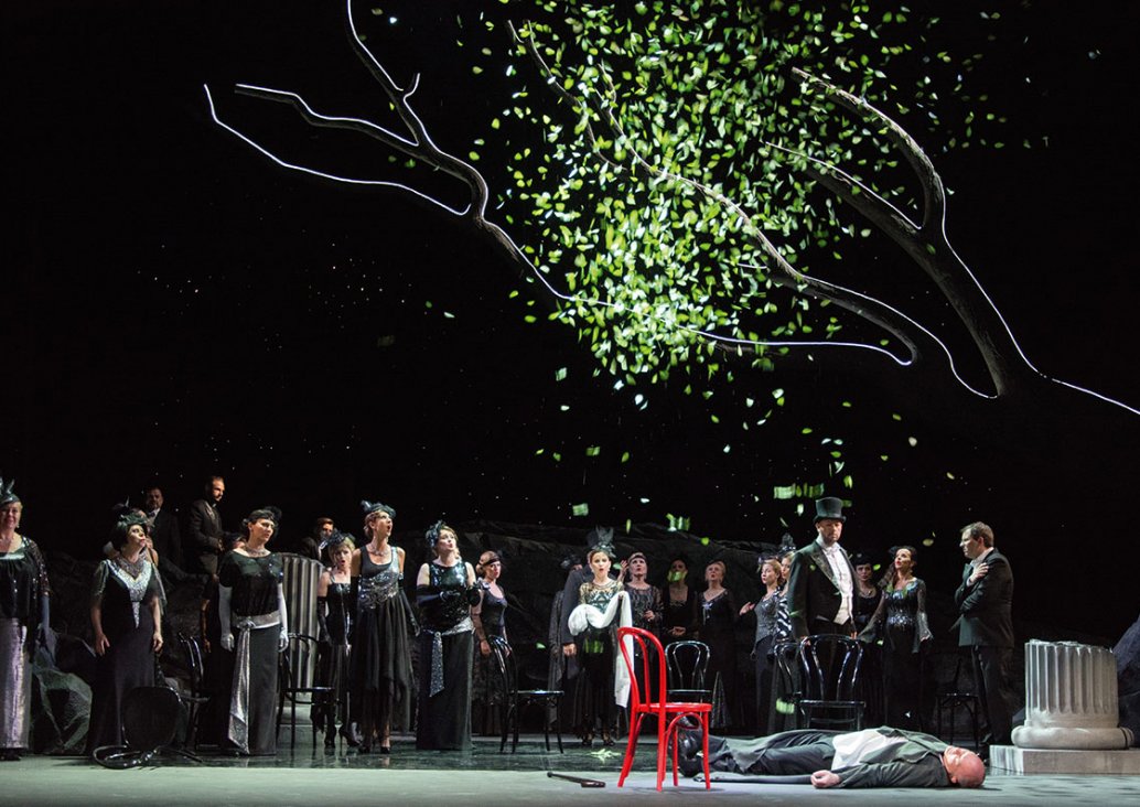 A group scene, a red chair in the center, a soloist lying next to it on the floor. Above it there is a tree with leaves falling from it, in the right year a fragment of a white column. In the background, characters in dark evening dresses and black chairs