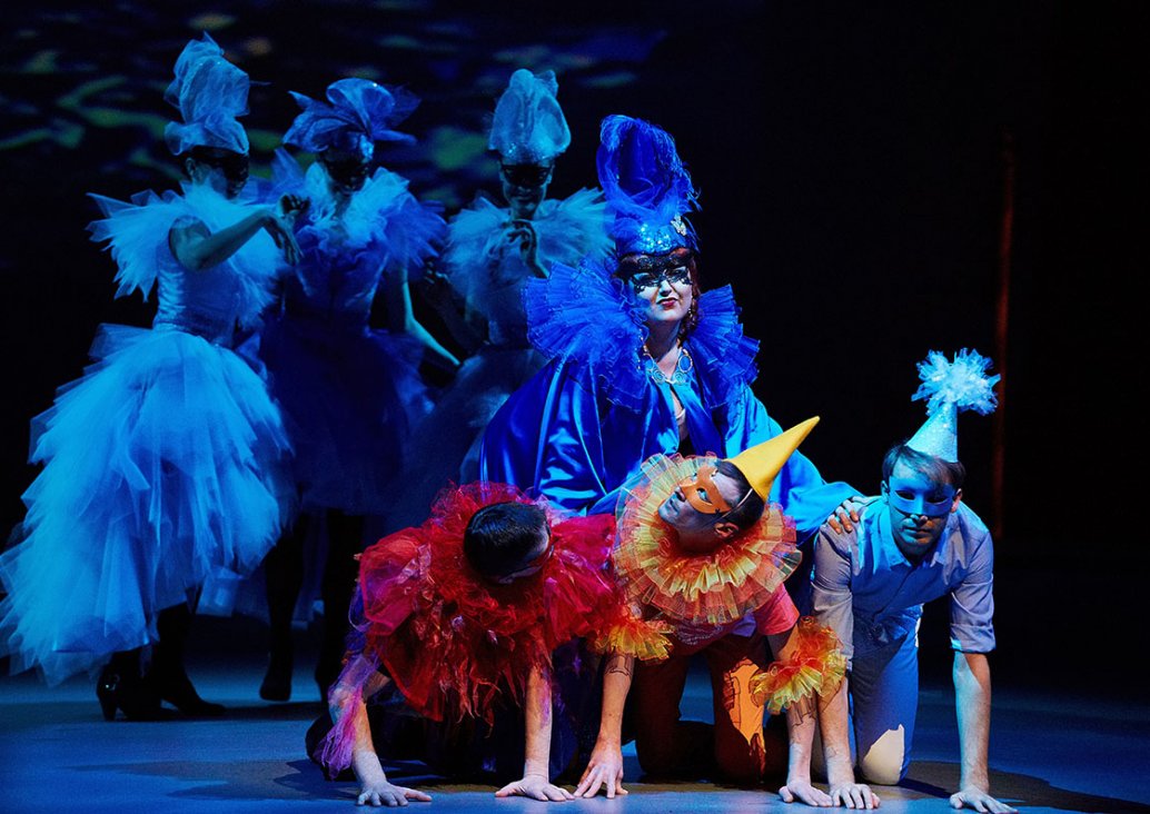 Scena grupowa, na pierwszym planie solistka w niebieskim stroju i masce podpiera się rękami o trzy postaci w pozycji klęku podpartego. W tle trzy postaci w karnawałowych sukniach i maskach.