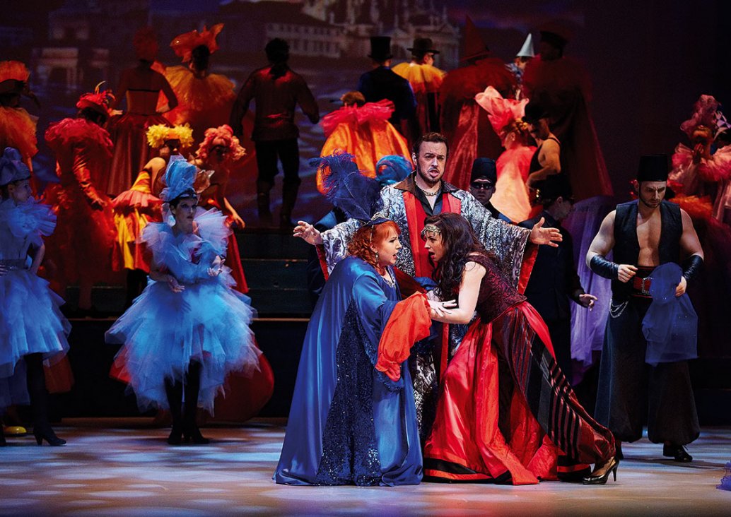 The group scene, with three soloists in the foreground, two soloists leaning towards each other and holding hands in their elbows, behind them the soloist with his arms spread out. Other characters in the background. All dressed in colorful, carnival cost