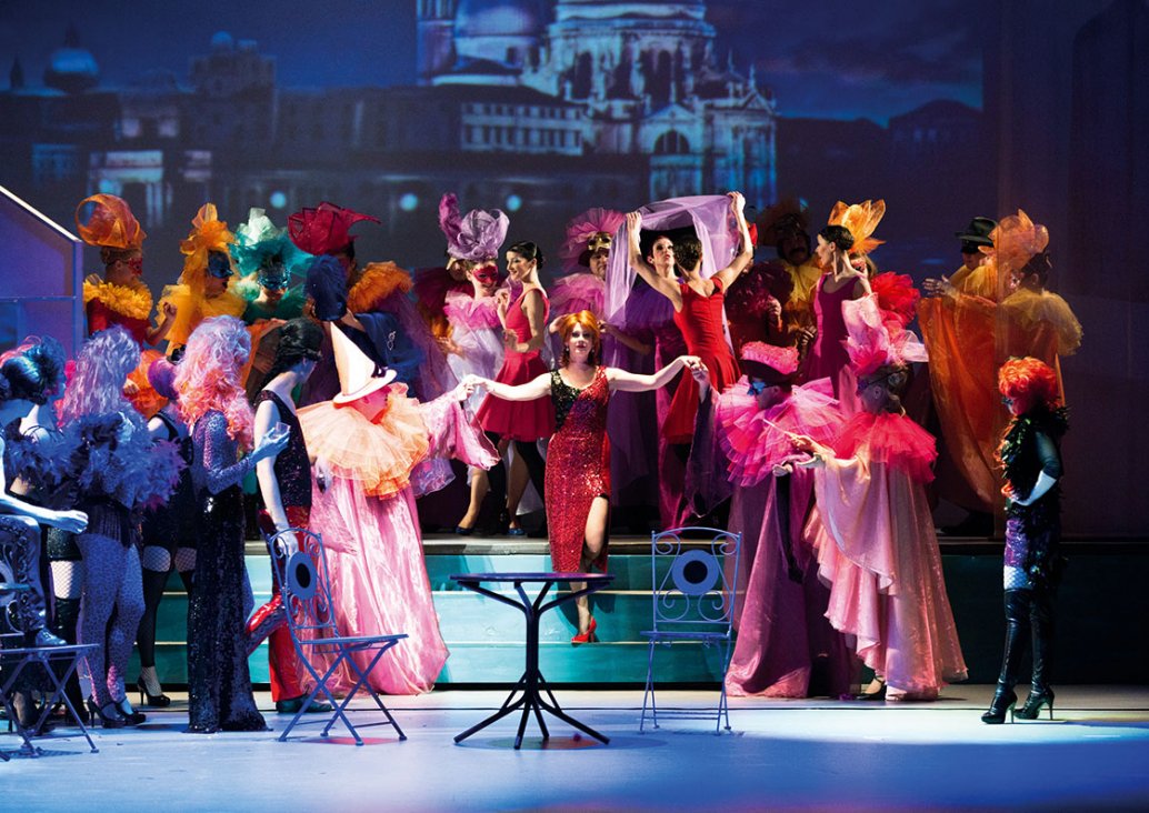 A group scene, in the middle of the three-stage staircase the soloist in a shiny red dress is holding two other characters by her hands. Everyone around the soloist dressed in colorful carnival costumes.