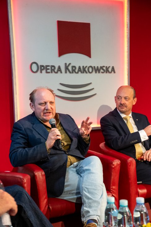 Brodaty mężczyzna w granatowej marynarce opowiada o czymś do mikrofonu gestykulując przy tym. Obok siedzi mężczyzna w garniturze i patrzy na niego.