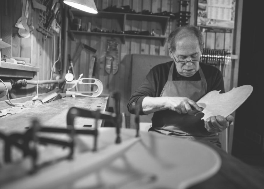 Jan Pawlikowski, ubrany w fartuch roboczy, pracuje w swoim zakładzie lutniczym nad spodnią częścią skrzypiec. Wnętrze jest oświetlone punktowo za pomocą lampki. W tle widać narzędzia lutnicze powieszone na ścianie.