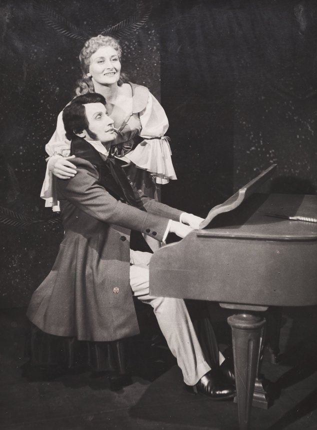 Zdjęcie ze spektaklu "Opowieści Hoffmanna". Kazimierz Pustelak (Hoffmann)gra na fortepianie, zwrócony bokiem do patrzącego. Za nim stoi Celina Kurpińska (Antonia), obejmuje go za ramiona. Oboje patrzą w dal rozmarzonym wzrokiem.