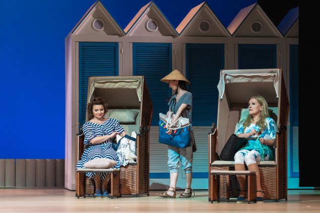 Kolorowa fotografia ze spektaklu "Cosi fan tutte". Scena na plaży. Dwie kobiety w niebiesko-białych strojach relaksują się, siedząc w koszach plażowych. Pomiędzy nimi stoi kobieta w trójkątnym azjatyckim kapeluszu, trzymająca dużą torbę. W tle niebieskie 