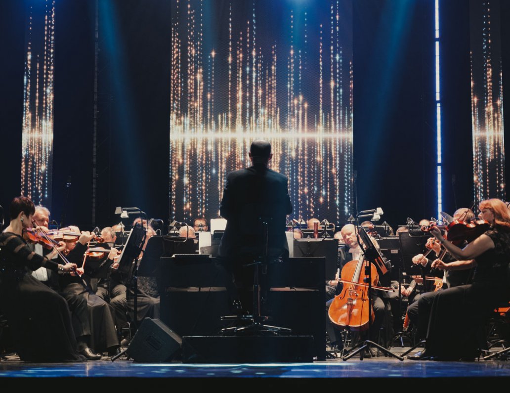 Na środku zdjęcia siedzi odwrócony plecami dyrygent, dookoła niego jest orkiestra. Nad głową dyrygenta jest poziomy rozbłysk światła